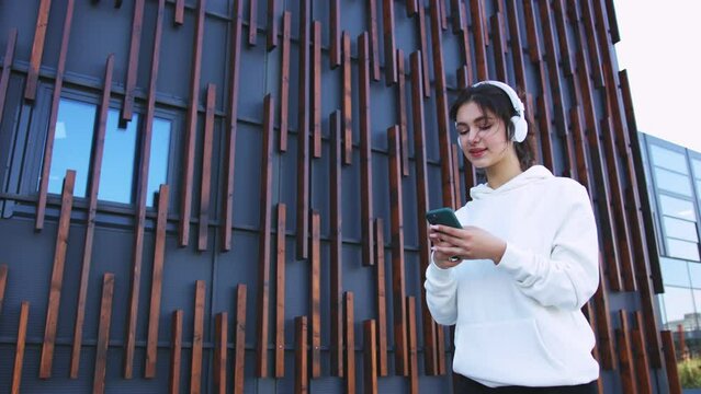 Pretty stylish young caucasian woman in sportswear and headphones walking the street, listening to music and texting mesage on smartphone. Female messaging online on mobile phone. Outdoor.