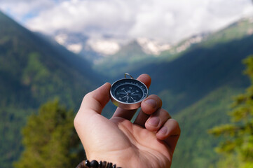 A hand with a compass against the backdrop of epic snow-capped mountains with clouds and a forest...