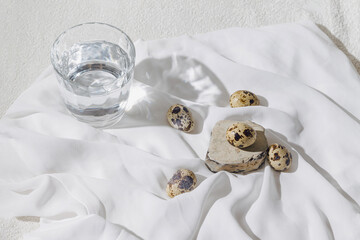 Creative layout made of quail eegs and glass of water on white fabric with sunlit and shadow. Healthy food ingredient concept. Natural easter background