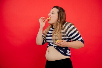 Overweight adipose woman with naked belly in striped clothes holding and eating potato chips....