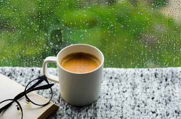 A cup of hot coffee with book and spectacles on table in morning with rain drop on window and green...