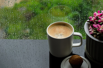A cup of hot coffee with small cake on table in morning with rain drop on window and green background. Stay home and relaxing concept.