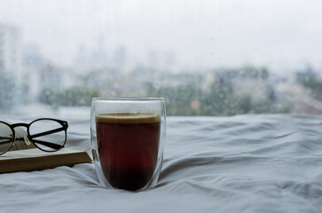 A glass of hot coffee with book and spectacles on bed in morning with rain drop on window. Stay...