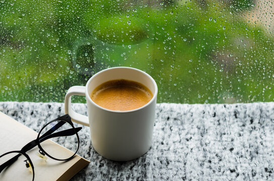 A cup of hot coffee with book and spectacles on table in morning with rain drop on window and green background. Stay home and relaxing concept. - Powered by Adobe