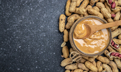 peanut butter or peanut paste in an open jar and peanuts in the peel scattered on a dark background, banner, menu, recipe place for text, top view