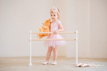 Cute blue-eyed baby ballerina in pink, standing near the ballet barre with a teddy bear in her...