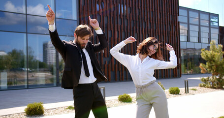 Couple of Caucasian young stylish businessman in suit and tie and businesswoman in glasses dancing and having fun outdoor. Successful in business man and woman doing dance moves and celebrating. - obrazy, fototapety, plakaty