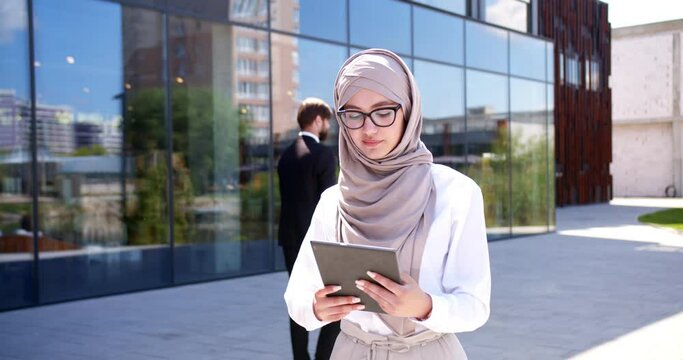 Young Arabian female in glasses and hijab walking the street in city business center and using tablet device. Beautiful Arab muslim woman rushing outdoor and tapping on computer. Arabic businesswoman.