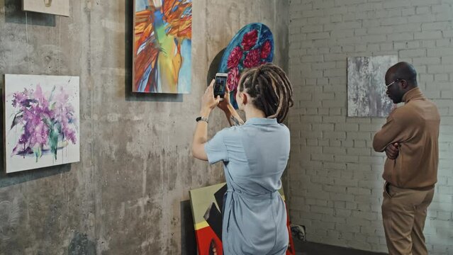 Rear view of young Caucasian woman with dreadlocks taking pictures with smartphone of paintings on wall in art gallery