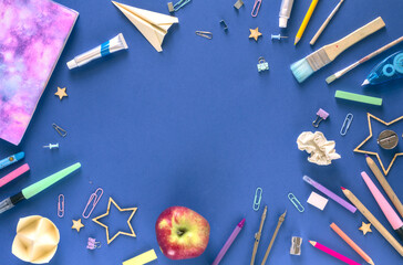 Back to school or education concept. Top view of various items in disorder: school supplies, origami, notebook, and an apple. A flat lay photo on navy blue background with copy space in the center.