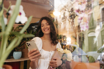 Beautiful caucasian young girl reads message on phone standing in flower shop. Brunette wearing dress buys bouquet as gift. Leisure, technology concept