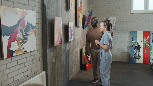 Medium long of Caucasian girl with dreadlocks and young Black man in eyeglasses talking and enjoying paintings on art exhibition at daytime