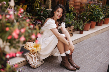 Cute european young girl looking at camera, sitting near flower market during day. Brunette wears white sundress and boots in summer. City life concept