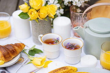 aromatic herbal tea in two cups, teapot, croissant and yellow roses, summer tea party in the garden