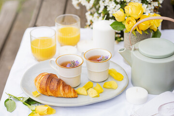 tea party in the garden, herbal tea, teapot, orange juice, croissant, yellow roses and white candles on a white tablecloth