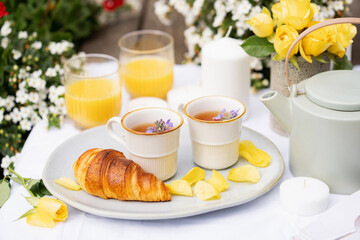 tea party in the garden for two, herbal tea, teapot, orange juice, croissant, yellow roses and white candles on a white tablecloth