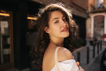 Close-up of fashionable young caucasian woman turns to camera on background of city. Brunette with wavy hair wears white blouse. Relaxation concept