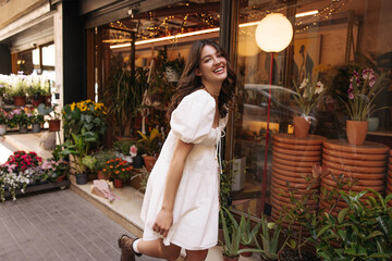 Pretty young caucasian woman laughing looking at camera walking in flower market. Brunette wears white sundress summer in warm weather. Mood, lifestyle, concept.