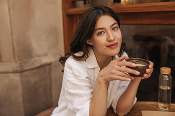 Pretty young caucasian woman looking at camera enjoying coffee in street cafe. Brunette with wavy...