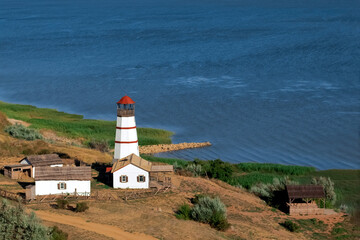 On the shore of the blue sea there is an old red-and-white lighthouse, next to the house of the lighthouse keeper, a barn, a pier made of stones, a road