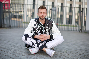 Obraz premium A handsome man in a white and black suit with a playful look. A guy with stubble and a model hairstyle in a trendy suit sits on the pavement.