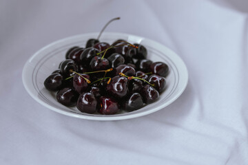 Fresh ripe dark black huge sweet cherries and water drops on white plate, light cloth background. Rustic organic fruits to eat.  Bio and eco berries for jam, juice, smoothie, desserts. Selective focus