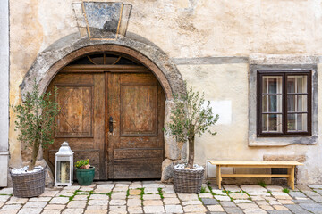 Fototapeta na wymiar Ein Altes Haus mit braunen Holztor und Fenster, davor eine Holzbank, Laterne und Blumentopf mit zwei kleine Olivenbäume