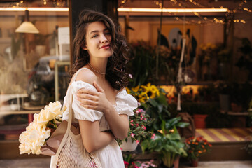 Cute young caucasian girl walks in flower shops outdoors during day. Brunette woman with wavy hair wears summer dress. Lifestyle concept