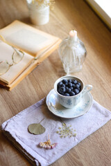 Obraz na płótnie Canvas Vintage porcelain cup filled with fresh blueberries, pressed flowers, open book, reading glasses and lit candle on the table. Hygge at home. Selective focus.