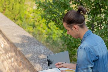 Young man on bun hairstyle looking down to his notebook enjoying leisure activity in the nature with laptop