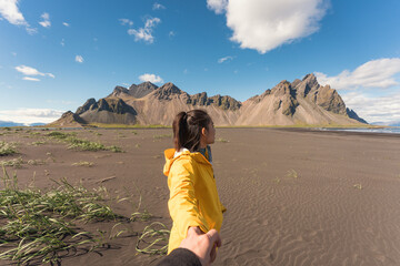 Fototapeta premium Young asian woman in yellow jacket holding hands with boyfriend on the beach with Vestrahorn mountain in viking village on Stokknes at Iceland