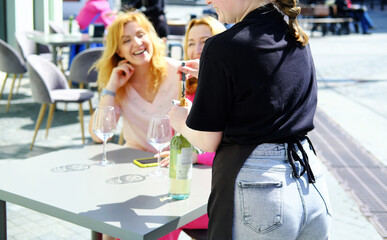 Two adult female friends are sitting at a cafe table on the street in summer and waiting for the waiter to open the wine