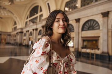 Obraz premium Beautiful young caucasian girl looks into camera spending time in large room. Brunette with wavy hair wears blouse summer. City life concept