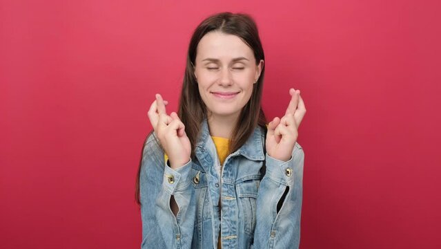 Cute young girl teen student in denim jacket and yellow backpack keep finger cross making with eyes closed, posing isolated on red background wall. Education in high school university college concept