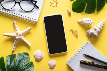 A background with a smartphone, glasses, a blank notepad and leaves and shells on a yellow background. Blogging in the summer at the workplace is a business. Copy space. Flat lay, top view