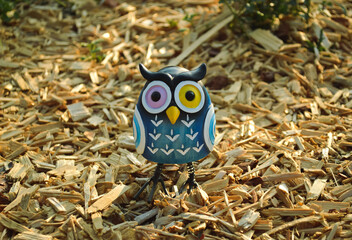 A toy of a surprised blue owl with multi-colored eyes close-up stands on wooden sawdust in the light of the evening sun