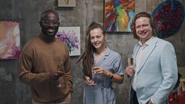 Medium portrait of cheerful Caucasian girl with dreadlocks, businessman and Black man holding glasses of champagne, standing in modern art gallery, looking and smiling on camera