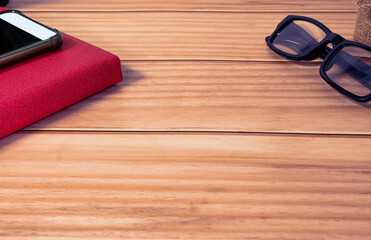 Cellphone, red note book, potted plant and reading glasses on wooden desk.
