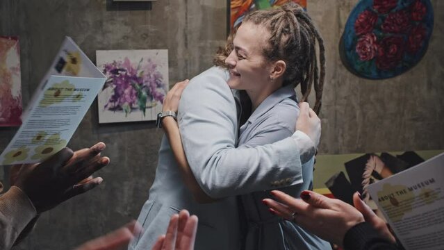 Medium slowmo of cheerful Caucasian girl with dreadlocks celebrating opening of her art exhibition, diverse people applauding and hugging artist