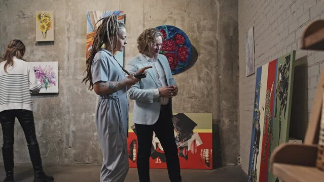 Medium long of blond-haired Caucasian man and girl with dreadlocks talking and looking at paintings in contemporary art gallery at daytime