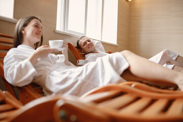 Cute couple in white bathrobes laying on a sunbeds indoors and drink a tea