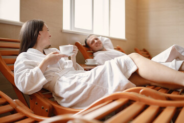 Cute couple in white bathrobes laying on a sunbeds indoors and drink a tea