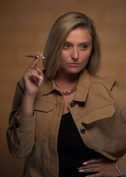 Portrait of a young, beautiful girl with blue eyes and blonde hair in a beige jacket. She looks sternly and smokes a cigarette. She is wearing an expensive watch, a gold chain and rings.