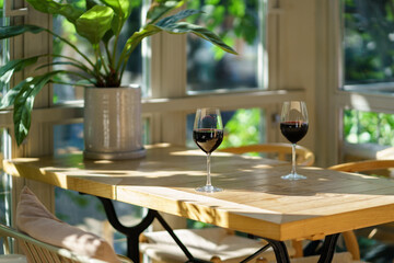 Cozy interior of terrace of summer cafe. Natural design soft light with shadow. Two trendy wineglasses of vintage red wine stand on wooden table in glare of sun. Soft cushions on chair for comfort.
