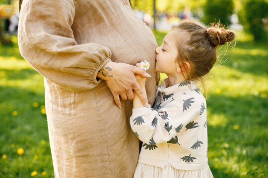 Cropped photo of a little girl kissing pregnant belly of mother in park