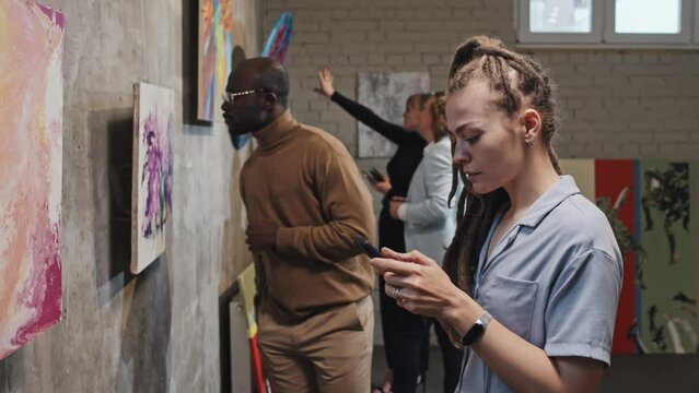 Side view of Caucasian girl with dreadlocks taking pictures with smartphone of paintings on exhibition at daytime, visitors walking around