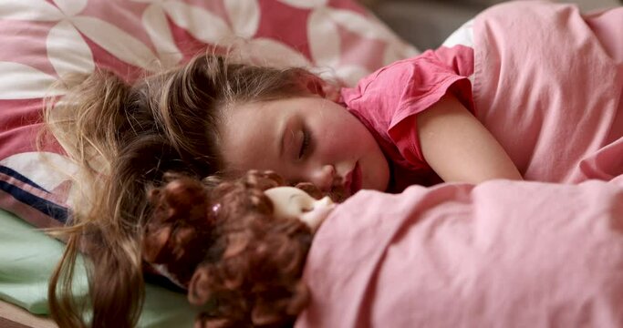 Little cute girl wakes up in the morning. Face close-up child opens eyes lying on pink bed linen next to toy at home