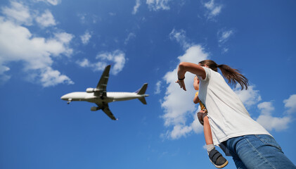 Bottom view of woman with toddler waving hand to landing commercial airplane in the sky. Lifestyle and travel concept.