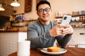 Adult asian man using cellphone while having breakfast in cafe indoors