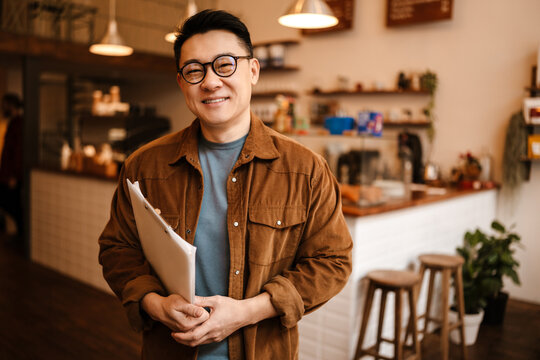 Adult asian man smiling and holding paper documents in cafe indoors